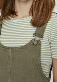 Salopette vert olive avec bretelles réglables et ferrures métalliques, portée sur une chemise rayée vert clair et blanc. Tissu texturé.