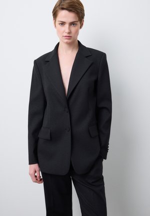 Schwarzer taillierter Blazer mit Halbschalrevers, zwei Knöpfen und drei Taschen, aus strukturiertem Stoff gefertigt. Kombiniert mit passenden schwarzen Hosen.