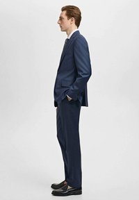 Navy suit with a tailored fit, single-breasted jacket, slim trousers, and black loafers. White shirt and patterned tie complete the look.