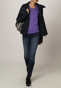Chaqueta de punto azul marino, camiseta morada, jeans skinny azul oscuro y zapatillas negras. El modelo lleva una bolsa con un diseño en blanco y negro.