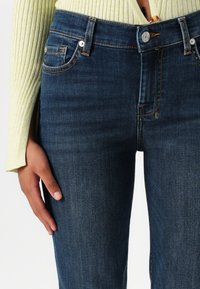 Dark blue slim-fit jeans with visible stitching, front pockets, and a button closure, displayed alongside a textured light yellow top.