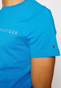 T-shirt in cotone blu con scollatura rotonda e maniche corte, decorata con il logo "HILFIGER" in grigio chiaro sul petto e un dettaglio con la bandiera sulla manica.