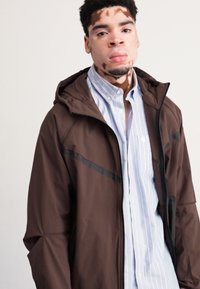 Brown hooded jacket with a smooth texture and black accent lines, layered over a light blue striped shirt.