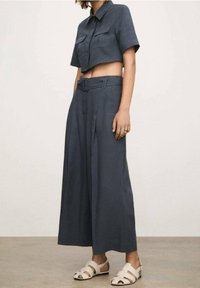 Camiseta gris corta con mangas cortas y dos bolsillos frontales, combinada con pantalones grises de pierna ancha que cuentan con un cinturón. Sandalias blancas con tiras.