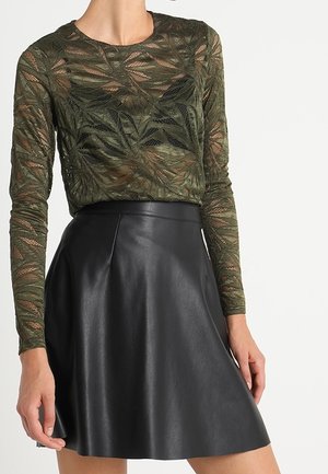 Frau trägt ein durchsichtiges grünes, mit Blättern gemustertes Langarmoberteil kombiniert mit einem hoch taillierten schwarzen Lederrock in A-Linien-Form.