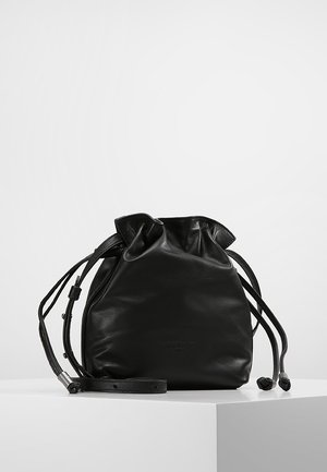Sac seau en cuir noir avec cordon de serrage et bandoulière ajustable, exposé sur une surface blanche avec fond gris.