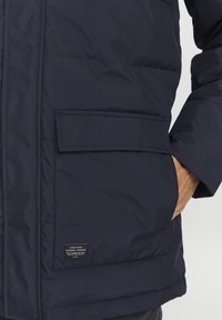 Marineblå pufferjakke med strukturert finish, som har en stor frontlomme og et merke på nedre venstre side.