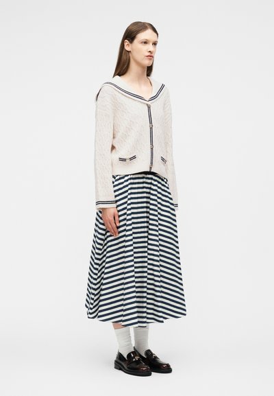 Kvinde iført en cremefarvet kabelstrikket cardigan med sømandsskrave, navyblå og hvide stribede plisserede midi-nederdel, hvide strømper og sorte loafers.