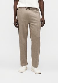 Ανοιχτό καφέ, ελαφρύς παντελόνι φόρμας με ελαστική μέση και κορδόνι. Έχει ευθεία γραμμή ποδιού και πλευρικές ραφές. Φοριέται με λευκά αθλητικά παπούτσια.