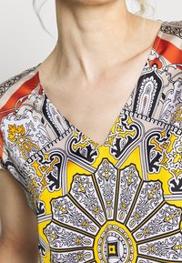 Haut à col en V à motifs avec des teintes vibrantes de jaune, noir et rouge ; motifs floraux et géométriques complexes sur un tissu léger.