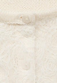 Krämfärgad stickad tröja med mjukt, texturerat garn; ribbad krage; och en stor, rund, genomskinlig knapp framtill.