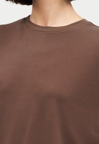 Κοντινό ενός ατόμου που φορά ένα απλό καφέ t-shirt με στρογγυλή λαιμόκοψη, δείχνοντας το λαιμό και το πάνω μέρος του στήθους.