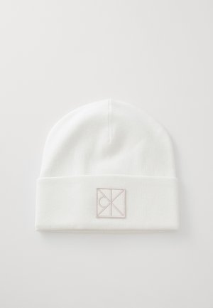 Witte gebreide beanie met een omgevouwen rand en een rechthoekig logo-label in licht taupe, met een gladde textuur en een afgeronde bovenkant.