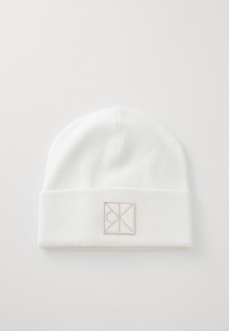 White knit beanie with a folded cuff featuring a rectangular logo patch in light taupe, smooth texture, and rounded top.