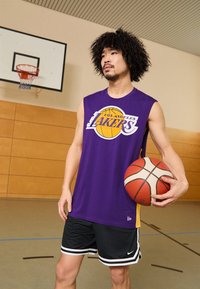 Vijolična brezrokavnik dresa Lakers z logotipom, črne kratke hlače z belimi črtami, drži košarko, postavljen v telovadnici.