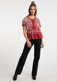 Bluse mit roten und weißen Mustern, Puffärmeln und gerafftem Bund. Kombiniert mit schwarzen, ausgestellten Hosen und spitzen schwarzen Stiefeln.