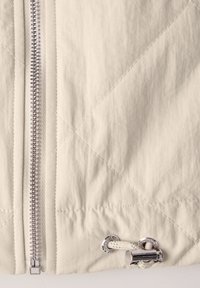 Veste beige matelassée avec une fermeture éclair argentée et des accessoires en métal poli. Se resserre avec un cordon et un accent métallique à l'ourlet.