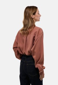 Blouse couleur rouille en tissu lisse avec une coupe décontractée, épaules tombantes et poignets à boutons, portée avec un jean en denim foncé.