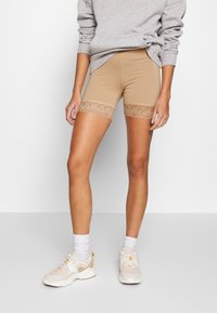 Béžové cyklistické šortky s dekorativní krajkovou vsadkou, vyrobené z pružného materiálu. Modelka má na sobě šedou mikinu a béžové sportovní boty s bílými ponožkami.