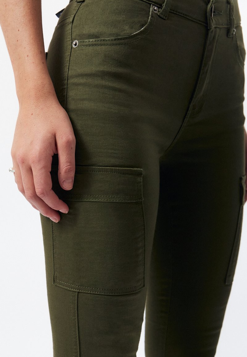 Pantalones cargo verde oliva con una textura suave, que cuentan con un bolsillo lateral y acentos de metal. Vista en primer plano de la tela y el ajuste.