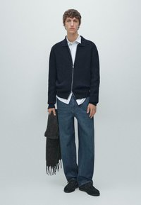 Giovane uomo con cardigan blu navy con zip sopra una camicia bianca, jeans blu larghi, scarpe nere, che tiene in mano destra una sciarpa grigia scura con frange.