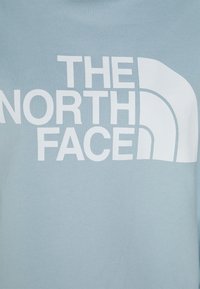 Ljusblå sweatshirt med en stor vit logotyp med texten "THE NORTH FACE" och ett distinkt böjt designelement. Mjuk tygtextur.