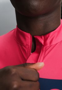 Persona de piel oscura abrochando la cremallera de una camisa de color rosa brillante con un cuello alto, mostrada en una vista en primer plano del cuello y la mano.