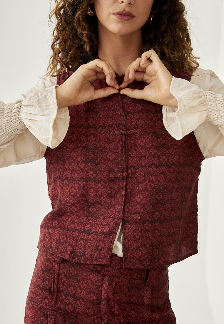 Chaleco con estampado floral rojo, hecho de una tela texturizada, que presenta un corte recto y cierres de nudos tradicionales, llevado sobre una blusa clara.