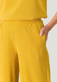 Pantalons en tissu texturé jaune avec une taille élastique et des poches latérales. Le design froissé ajoute un intérêt visuel et de la profondeur.