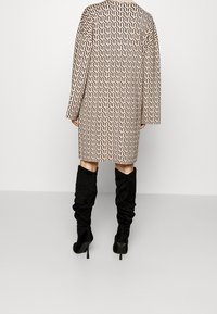 Cappotto lungo con un motivo geometrico in beige e marrone, dotato di maniche lunghe. Abbinato a stivali neri al ginocchio. Texture liscia.