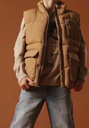 Henkilö, jolla on yllään vaaleanruskea toppaliivi beigenvärisen, mustalla grafiikalla varustetun neuleen päällä, siniset farkut, seisomassa ruskean taustan edessä.