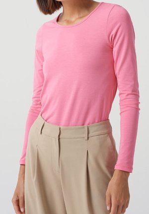 Femme portant une chemise rose ajustée à manches longues rentrée dans un pantalon beige plissé taille haute avec passants de ceinture, debout devant un fond uni.