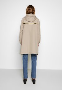 Beżowy płaszcz przeciwdeszczowy z kapturem, z żółtym wykończeniem z tyłu, w zestawieniu z granatowymi dżinsami i czarnymi botkami na obcasie. Widok z tyłu.