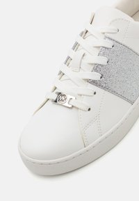 Sneaker bianco con tomaia in pelle liscia e fascia glitter argento. Presenta lacci bianchi e un accento logo argento lucido sulla linguetta.