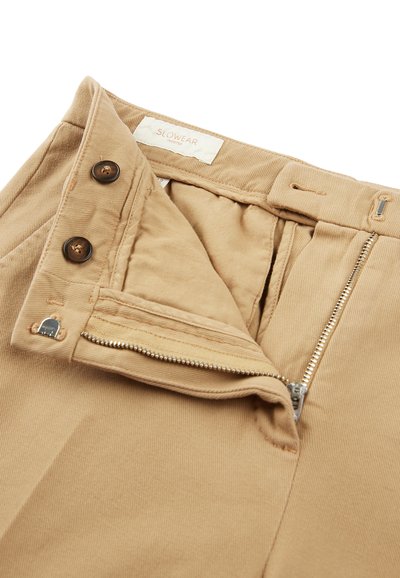 Beżowe spodnie z korduroy z zapięciem na guzik, metalowym zamkiem błyskawicznym i gładką podszewką; zawierają dwa brązowe guziki i etykietę marki.