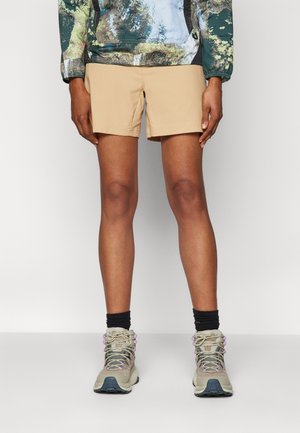 Pantaloni corti beige realizzati in tessuto leggero, abbinati a scarponi da trekking grigi e calze nere. Lo sfondo è bianco semplice.