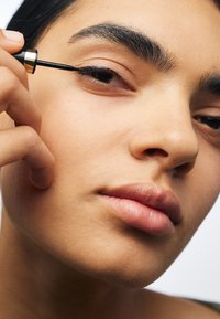 Svart eyelinerborste appliceras på den övre ögonfransen hos en person med slät, ljus hy, fylliga läppar och mörka ögonbryn, mot en neutral bakgrund.