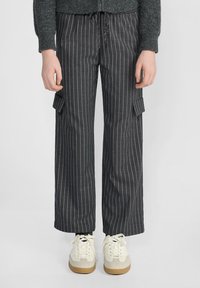 Personne portant un pantalon gris foncé à rayures avec des poches cargo et des baskets blanches, se tenant devant un fond uni.