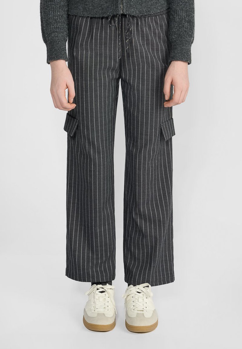 Personne portant un pantalon gris foncé à rayures avec des poches cargo et des baskets blanches, se tenant devant un fond uni.