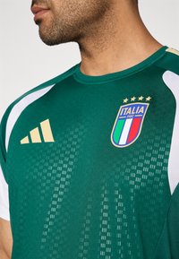 Camisola atlética verde com detalhes brancos, apresentando um decote redondo, textura suave, o logótipo da seleção italiana e três riscas douradas no ombro.