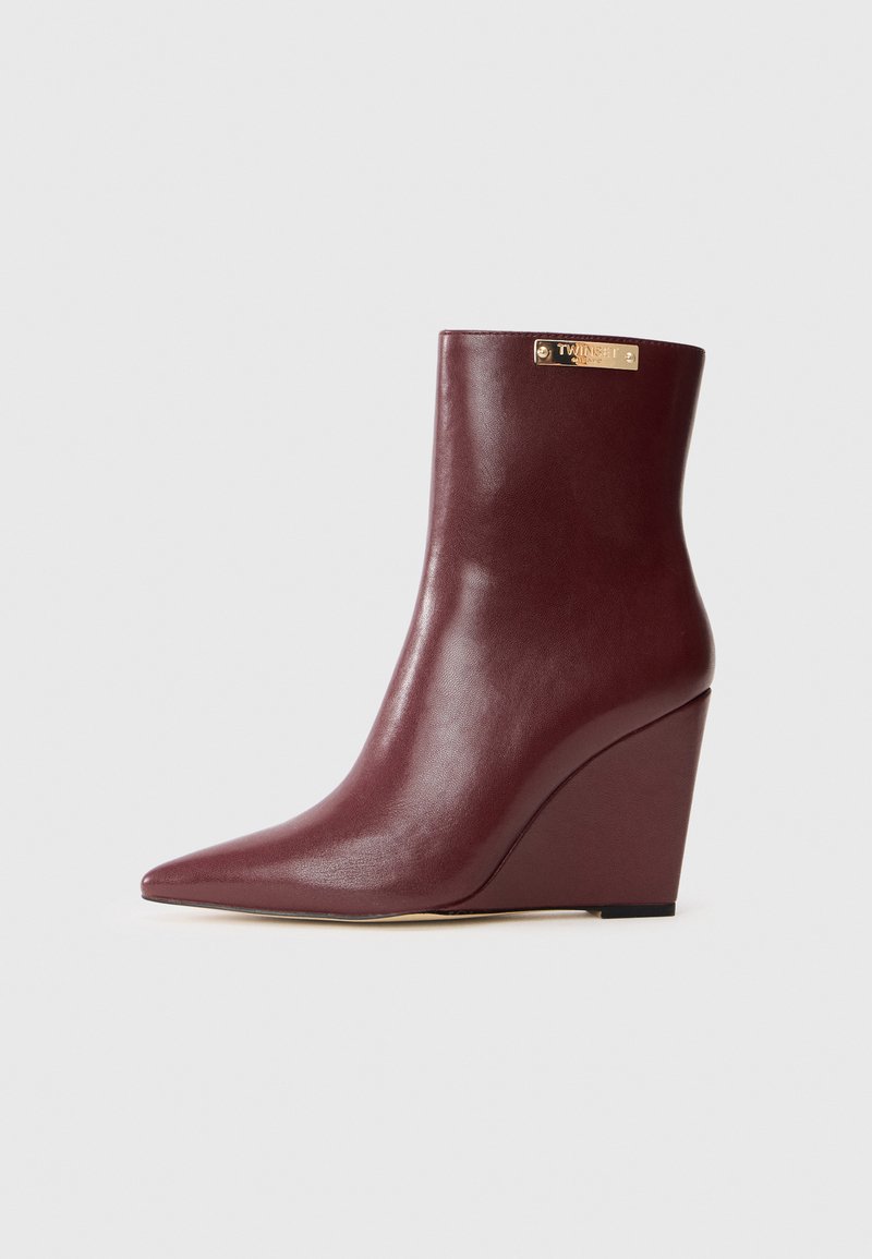 Bottine à talon compensé en cuir bourgogne avec un bout pointu, texture lisse et plaque logo dorée en haut. Silhouette élégante et moderne.