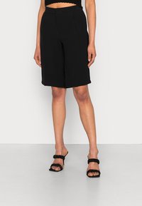 Shorts neri su misura con una vestibilità rilassata, caratterizzati da tasche e una texture liscia. Abbinati a sandali neri senza chiusura con cinghie arricciate.
