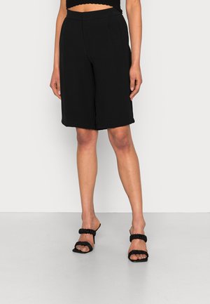 Shorts noirs ajustés avec une coupe décontractée, dotés de poches et d'une texture lisse. Associés à des sandales noires à enfiler avec des brides volantées.