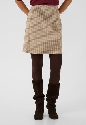 Femme portant une jupe beige à motif pied-de-poule, des collants marron foncé et des bottes en daim marron foncé et amples, debout les mains dans le dos.