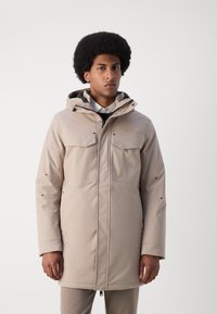 Parka impermeable beige con capucha, dos bolsillos en el pecho y bolsillos laterales con cremallera. Presenta una textura suave y elementos ajustables para un mejor ajuste.