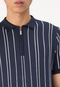 Polo tričko s námořnickými a bílými pruhy se zipovým límcem. Vyrobeno z měkkého materiálu, má krátké rukávy a přiléhavý střih.