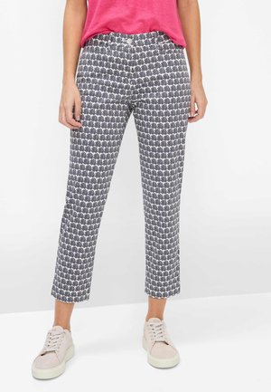 Person iført kapribukser med sort og hvidt bladmønster, lyserød top og lyserøde sneakers, stående på hvidt gulv.