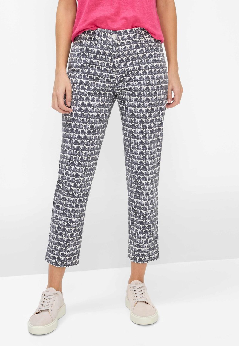 Person iført kapribukser med sort og hvidt bladmønster, lyserød top og lyserøde sneakers, stående på hvidt gulv.