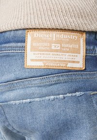 Jean en denim bleu clair avec une étiquette en cuir marron clair Diesel Industry cousue au-dessus de la poche arrière sous un pull en maille beige.