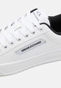 Biele tenisky s hladkým syntetickým zvrškom, okrúhlou špičkou, čiernymi akcentmi a plochými bielymi šnúrkami. Na boku sú s brandingom "Armani Exchange".